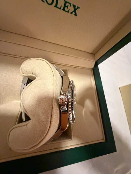 A Rolex watch displayed inside an open cream-colored Rolex presentation box with a cream cushion. The watch has a silver-toned metal case and a gold-toned bracelet.